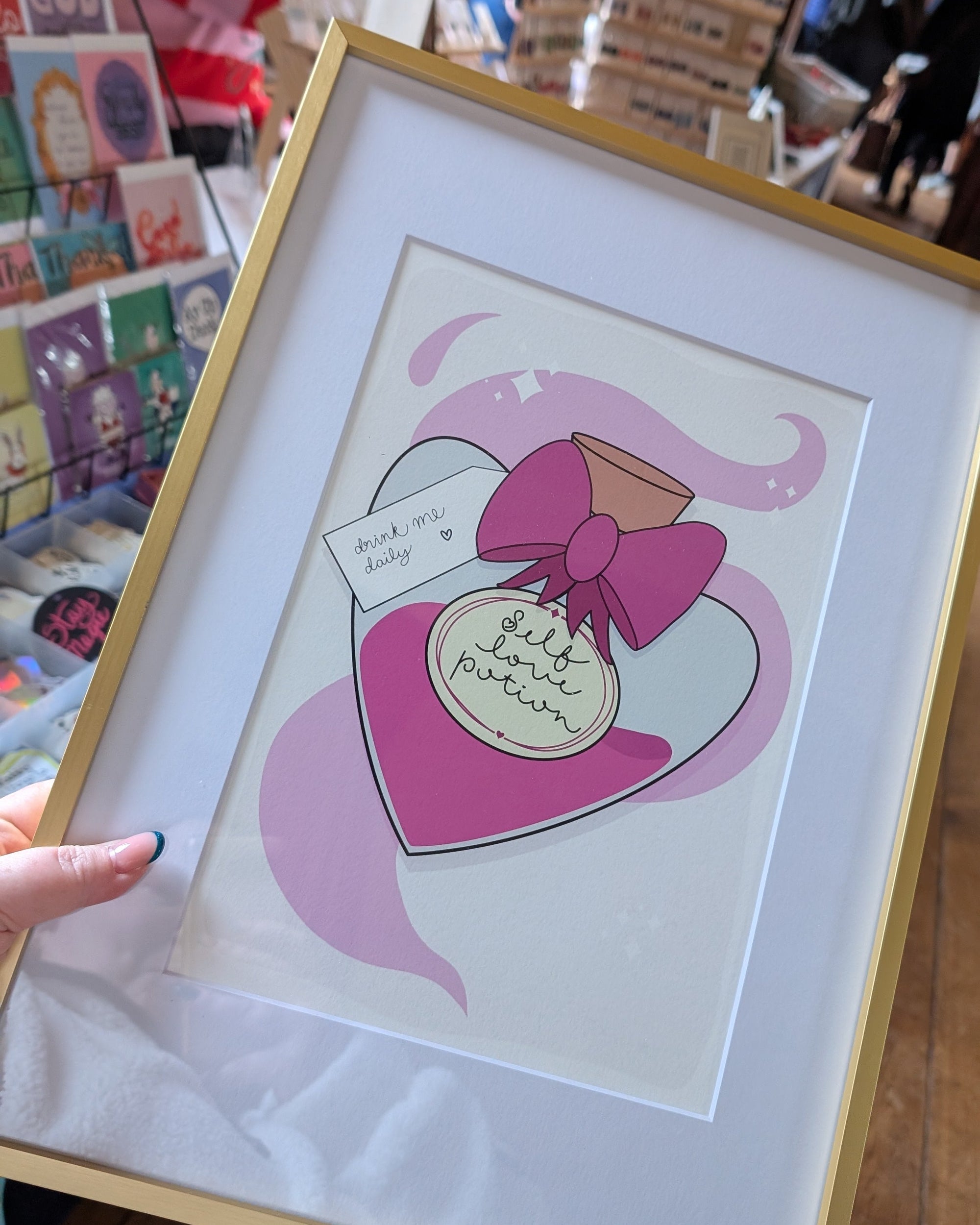 Framed artwork of a pink heart with a bow and text, held by a person.
