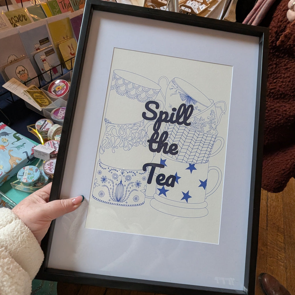 Framed artwork with 'Spill the Tea' text held by a person in a store setting.