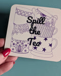 A hand with red nail polish holds a square white coaster featuring purple line art of teacups and the words 'Spill the Tea' in black script, set against a teal desk.