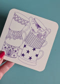 A hand with red nail polish holds a square white coaster featuring purple line-art illustrations of stacked teacups, set against a teal background.
