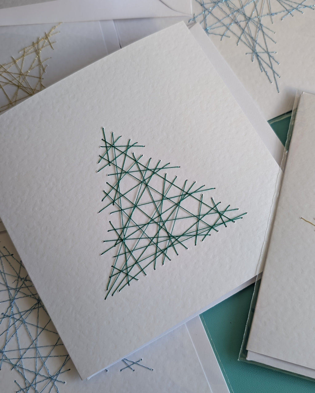 A stack of white textured paper cards featuring minimalist geometric line art of a Christmas tree and snowflakes in green and blue, arranged on a teal surface.