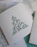 A stack of white textured paper cards featuring minimalist geometric line art of a Christmas tree and snowflakes in green and blue, arranged on a teal surface.