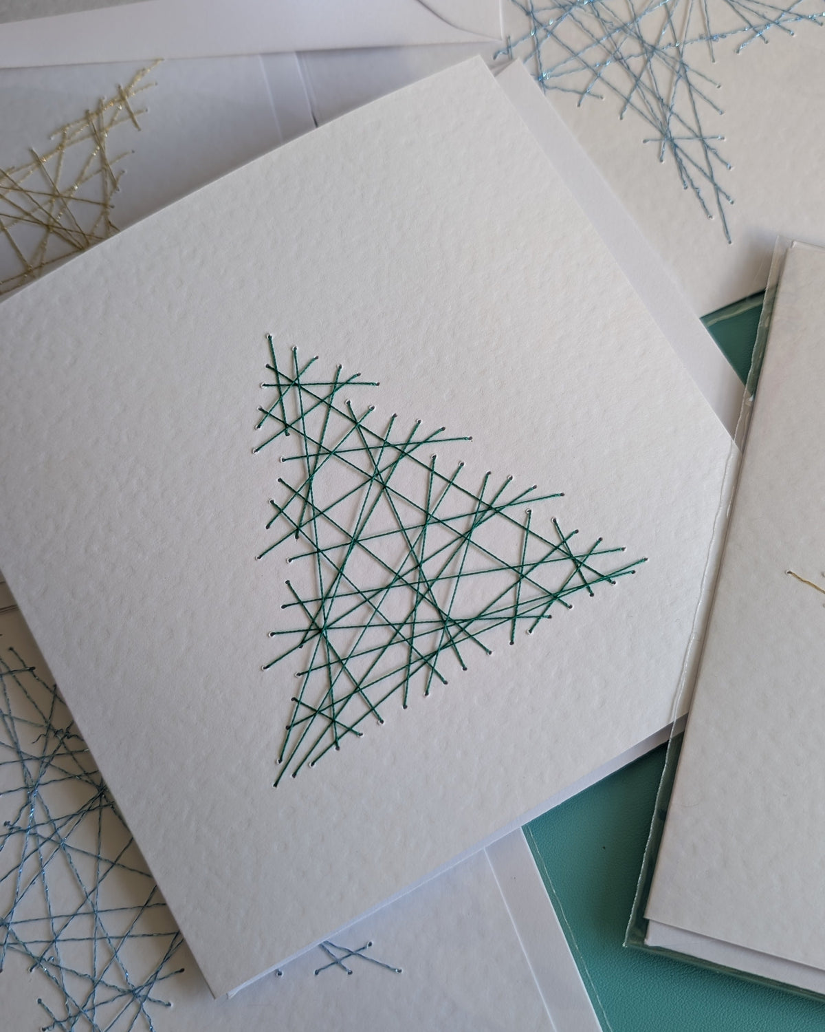 A stack of white textured paper cards featuring minimalist geometric line art of a Christmas tree and snowflakes in green and blue, arranged on a teal surface.