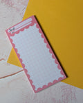 A pink notepad with a scalloped border and grid lines, placed diagonally on top of a yellow sheet over a textured light pink and white surface.