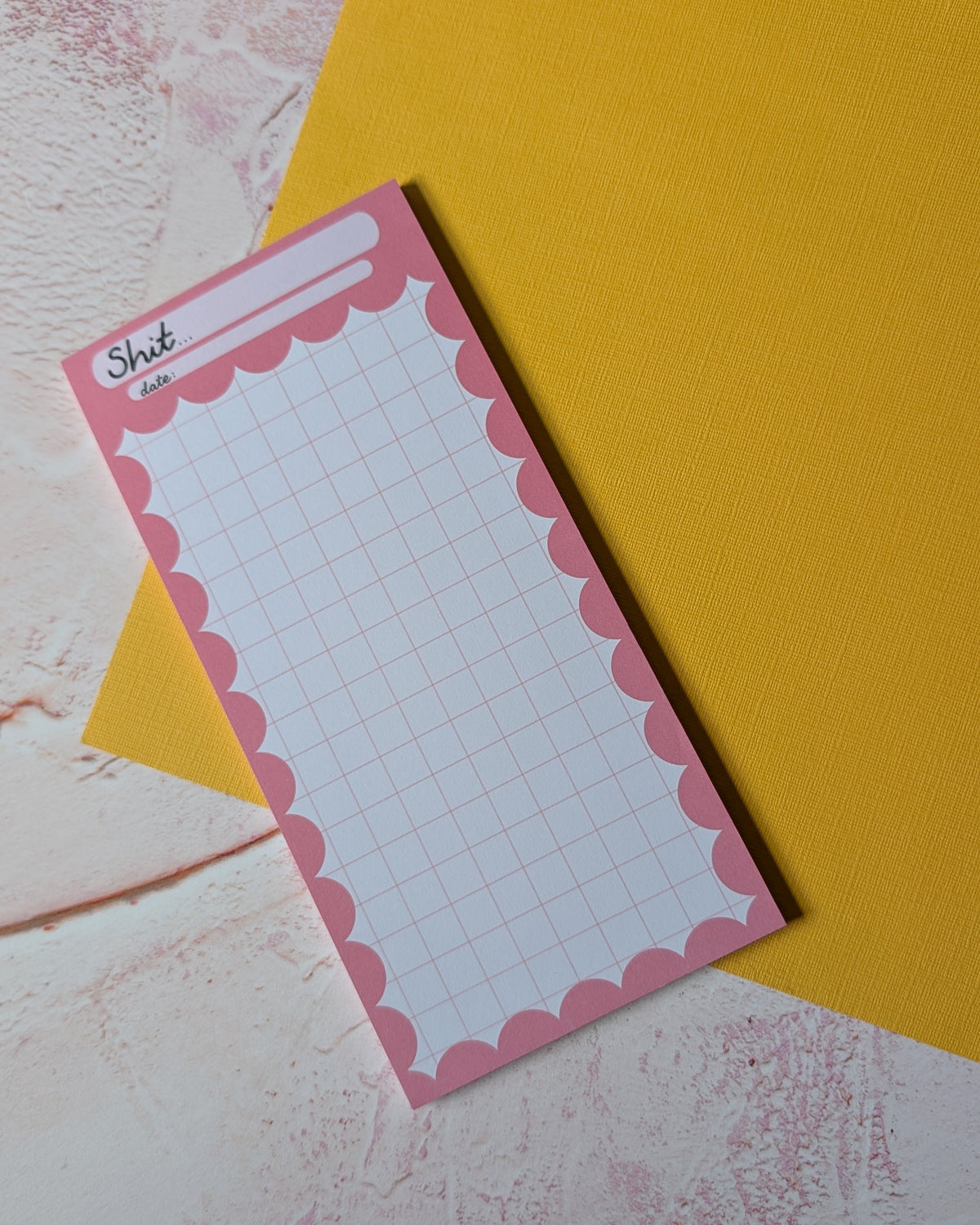 A pink notepad with a scalloped border and grid lines, placed diagonally on top of a yellow sheet over a textured light pink and white surface.