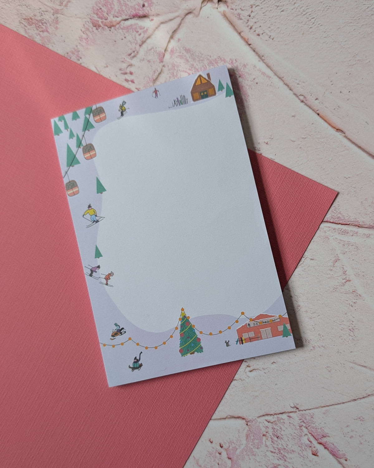 A rectangular white notepad with a whimsical illustrated border featuring a Christmas tree, house, bunting, and small figures, placed on top of a pink paper sheet over a textured white surface.