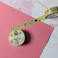 A roll of decorative washi tape with a yellow background and colorful cartoon illustrations, partially unrolled next to a pink sheet of paper on a textured white surface.