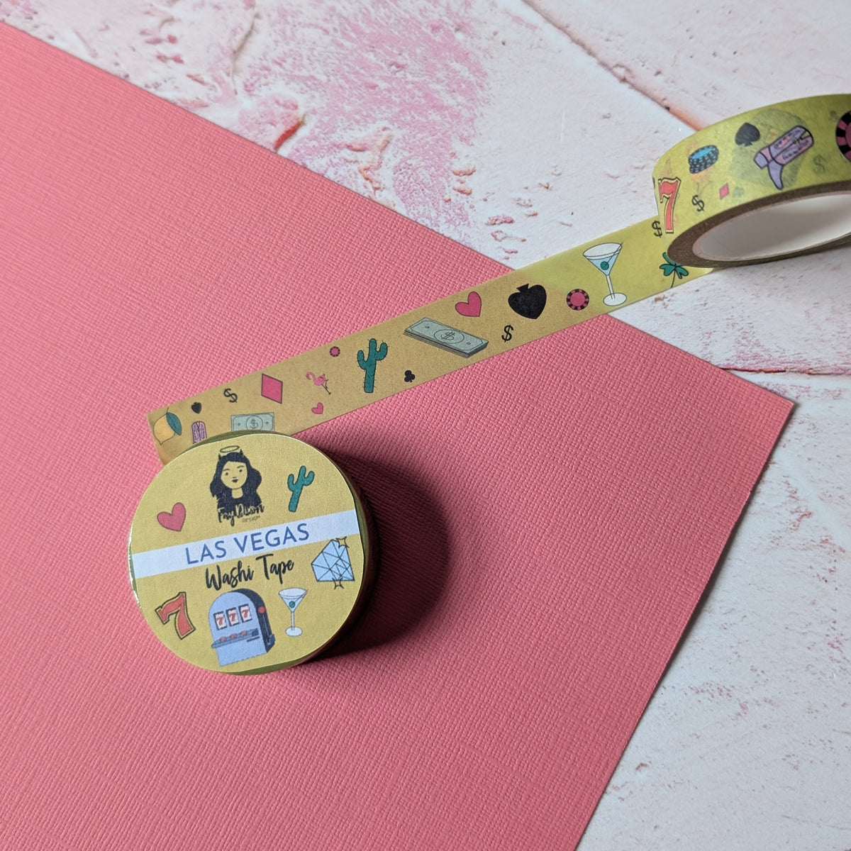 A roll of decorative washi tape with a yellow background and colorful cartoon illustrations, partially unrolled next to a pink sheet of paper on a textured white surface.