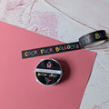 A roll of black washi tape with colorful text and a matching circular sticker are placed on a pink sheet over a textured white and pink background.