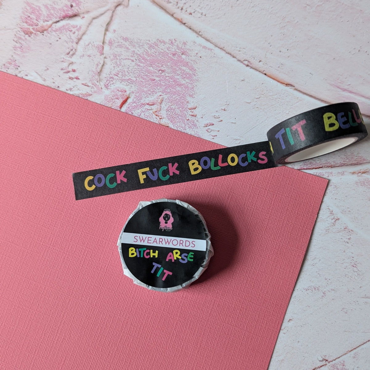 A roll of black washi tape with colorful text and a matching circular sticker are placed on a pink sheet over a textured white and pink background.
