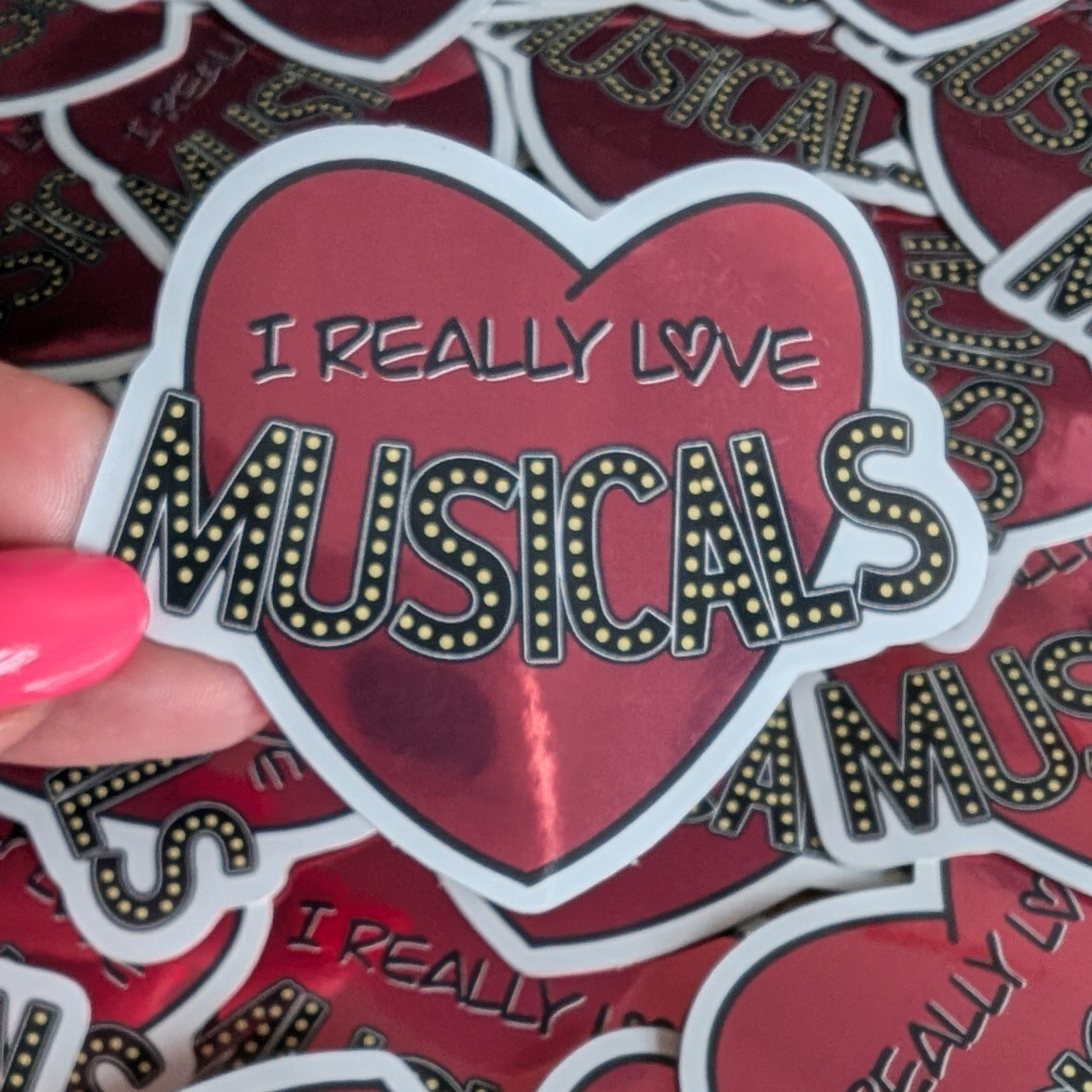 Heart-shaped sticker with 'I Really Love Musicals' text held over a pile of similar stickers.